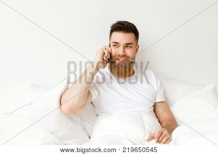Handsome young male with thick beard and musttache dressed in casual clothes sits on comfortable bed, has conversation with girlfriend, looks happily aside. People, communication and leisure concept
