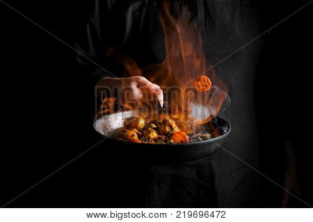 Chef cooking vegetables on a pan with fire flambe. Black background for copy text.