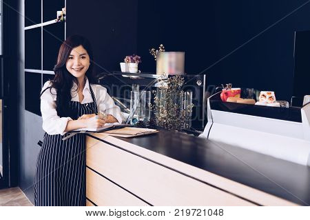 small business owner with notebook at counter in coffee shop. asian female barista wearing apron writing note at bar in cafe. food service restaurant entrepreneur concept.