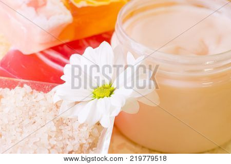 Facecare and bodycare concept. A close up of a container with smooth cream and bowl of pink bath salt, colorful soap bars, clean towels and a white flower. Space for a text or product display.