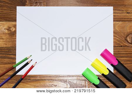 A white sheet of paper lay on a wooden table, near , pencils, markers, pens . The view from the top
