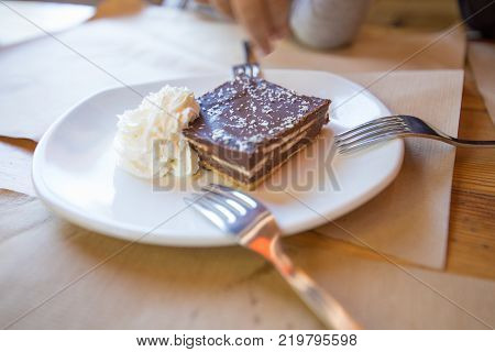 square piece of brown chocolate cake with cream on white ceramic dish with three forks for sharing on paper placemats and wooden table