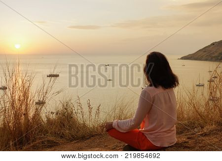 a Young girl sitting in yoga lotus meditation position in front to seaside on the rocks an watching the sun goes down in a golden hour. sunset over sea.