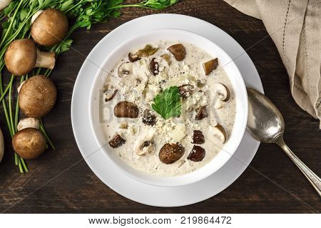 An overhead photo of a plate of a mushroom cream soup with champignons, cremini, fresh parsley, on a dark rustic background texture with a place for text