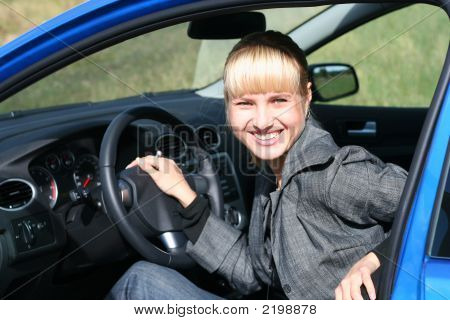 Young Blond Woman In A Blue Car. She Is Smiling Happy