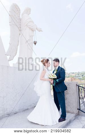 Portrait of a lovely couple honeymooned on a wedding day with a bouquet in hand against the background of an Orthodox Christian monument with angels. The concept of the Christian wedding in the church 