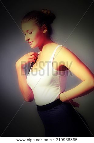A handsome girl is posing in a photo studio with a white background. She has a dramatic look. She is wearing a white shirt and grey sports skirt. With exposed shoulders. She has nice brown hair in bun
