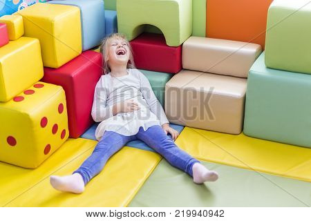 Little cute caucasian blond girl die laughing while playing with soft building blocks at indoor playground.
