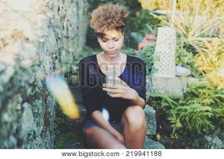 Young charming black female is aping and making faces while taking selfie on front camera of her smartphone in garden; African American girl is photographing herself on cellphone and making duckface