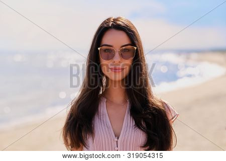 Portrait Of Beautiful Smiling Girl In Sunglasses And Pink Dress At Calm Seaside.