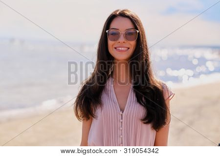 Portrait Of Beautiful Smiling Girl In Sunglasses And Pink Dress At Calm Seaside.