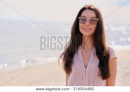 Portrait Of Beautiful Smiling Girl In Sunglasses And Pink Dress At Calm Seaside.