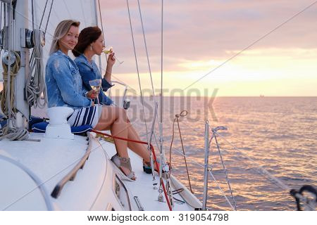 Two Joyful Women Are Celebraiting Good Summer Day At The Yacht While Watching Beautiful Sunset.