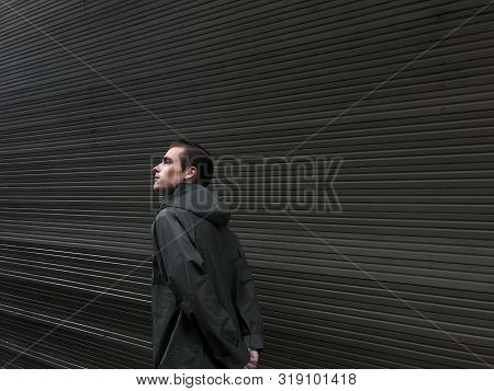 Handsome Young Man In A Raincoat Stands In Front Of A Wall, Garage Doors. Cope Space, Place For Text