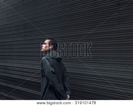 Handsome Young Man In A Raincoat Stands In Front Of A Wall, Garage Doors. Cope Space, Place For Text