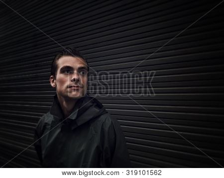 Handsome Young Man In A Raincoat Stands In Front Of A Wall, Garage Doors. Cope Space, Place For Text