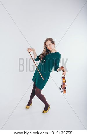 A Broken Doll In A Green Dress With A Violin And A Bow In Her Hands On A White Background. Tired Gir