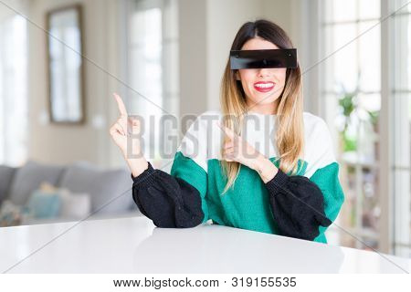 Young beautiful woman blindfold eyes with black censor band at home smiling and looking at the camera pointing with two hands and fingers to the side.
