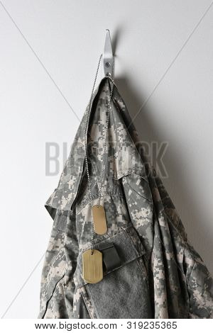 A set of military dog tags and camouflage field jacket hanging from a hook on a blank wall.