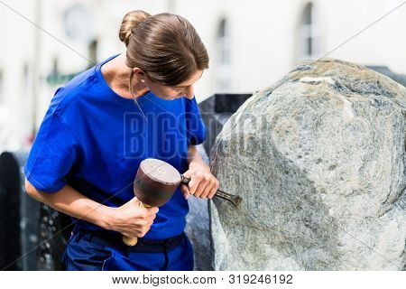 Female stonemason working on boulder with sledgehammer and iron