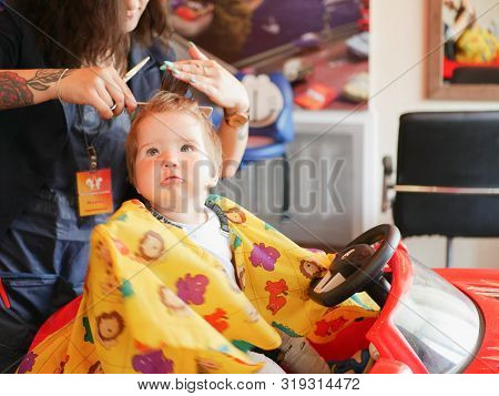 First Baby Haircut. Cute Blonde Toddler Is Happy To Be On A Haircut With A Professional Baby Hairdre
