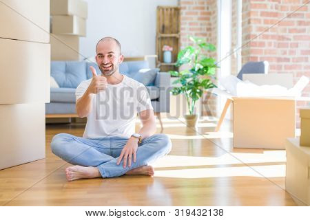 Young bald man sitting on the floor around cardboard boxes moving to a new home doing happy thumbs up gesture with hand. Approving expression looking at the camera with showing success.