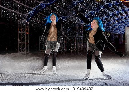 Two Identical Girls With Blut Hair Skating On The Ice Arena In The Evening City Square In Winter