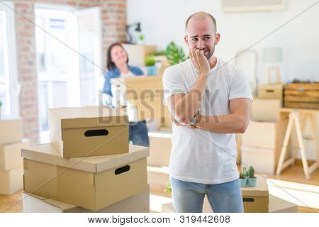 Young couple arround cardboard boxes moving to a new house, bald man standing at home looking stressed and nervous with hands on mouth biting nails. Anxiety problem.
