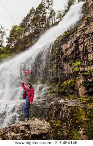 Tourist Woman With Norwegian Flag At Svandalsfossen In Norway, Powerful Waterfall In Norwegian Mount
