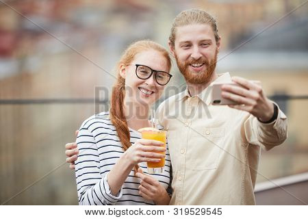 Young Man Embarcing Young Woman With Cocktail And They Smiling At Camera While Making Selfie Portrai