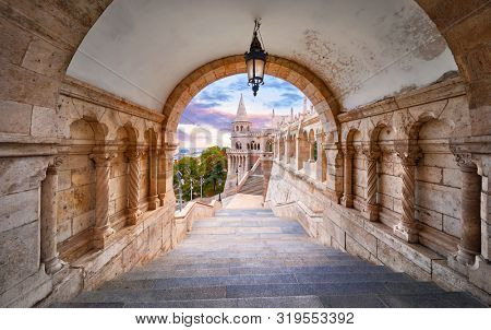 Budapest, Hungary. Ancient Fishermans Bastion castle. View at tower from stairs under arch along gallery with columns and street lantern. Picturesque evening sunset with clouds on sky. Famous.