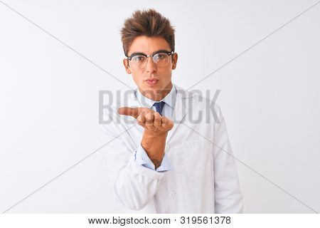 Young handsome sciencist man wearing glasses and coat over isolated white background looking at the camera blowing a kiss with hand on air being lovely and sexy. Love expression.
