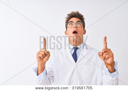 Young handsome sciencist man wearing glasses and coat over isolated white background amazed and surprised looking up and pointing with fingers and raised arms.
