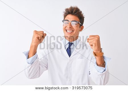 Young handsome sciencist man wearing glasses and coat over isolated white background very happy and excited doing winner gesture with arms raised, smiling and screaming for success. 