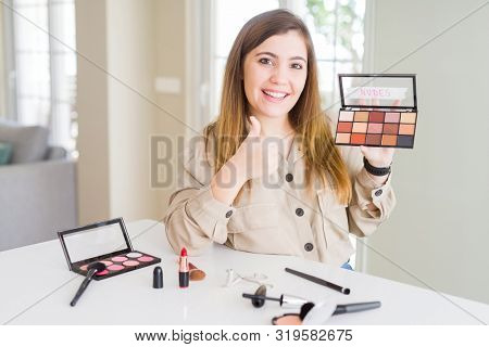 Beautiful young woman using make up cosmetics applying color from palete happy with big smile doing ok sign, thumb up with fingers, excellent sign