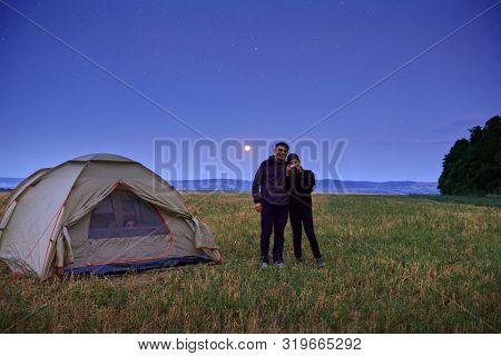 Family traveling and camping, twilight, posing near tent. Beautiful nature - field, forest, stars and moon. Father, son and girl sitting in tent. Boy taking photo by the camera.
