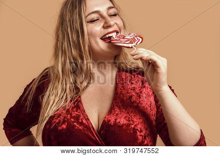 Mmm. Plus Size Happy Woman In Velour Dress Eating Red Heart-shaped Lollipop And Keeping Eyes Closed 
