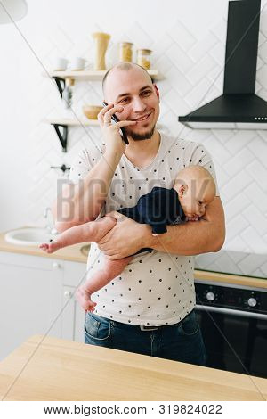 A Young Man In The Litchen Talking On Phone And Holding His Little Baby
