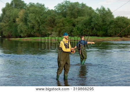 Two Fishermen Are Standing In The River, Wearing Rubber Boots. Men Throw Fishing Rods. Fishing Adven