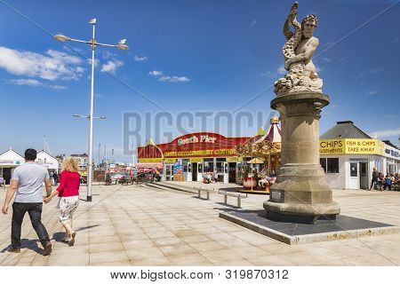 15 June 2019: Lowestoft, Suffolk, Uk - A Triton Statue On The Promenade, With South Pier, Amusements