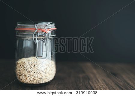 Close-up Shot Of Oat Flakes, Oatmeal In Weck Glass Jar On Rustic Wooden Table Against Dark Backgroun