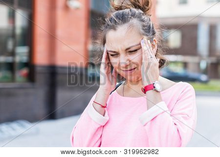 Young Caucasian Woman Winces Holding Her Head Suffering From A Headache On The Summer Street.