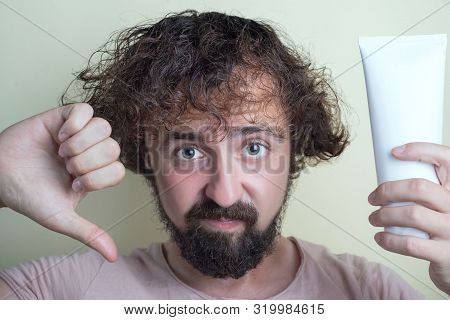 Portrait Of A Young Guy With Dirty Hair And Problematic Scalp. Holding A Bad Shampoo In Hand, Showin