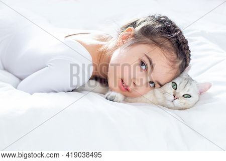 Portrait Of A Beautiful Little Girl In White Clothes, Lies On A Clean, White Bed With A White Britis