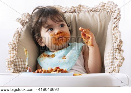 Adorable Little Child Funny Girl Eating Spaghetti With Spoon While Sitting In High-powered Chair At 