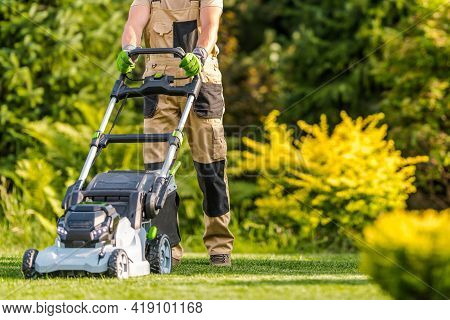 Caucasian Garden Worker In His 40s Cutting Grass Using Modern Electric Cordless Lawnmower In Large B