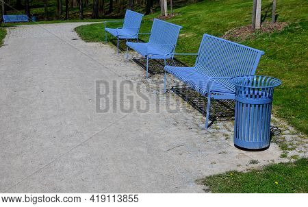 Park Benches In Light Blue Made Of Metal Strips Similar To A Lattice. Trash Can And Beautiful Lawn W