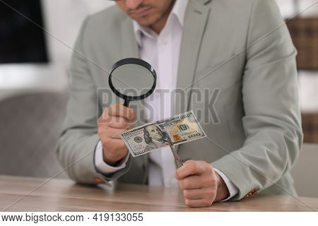 Expert Authenticating 100 Dollar Banknote With Magnifying Glass At Table In Office, Closeup. Fake Mo