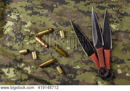 Black Throwing Knives And Casings On Camouflage Cloth. Three Special Knives With Red Handles Crossed