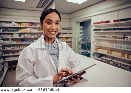 Portrait Of Smiling Young Female Pharmacist Wearing Labcoat Standing Behind Counter Using Digital Ta